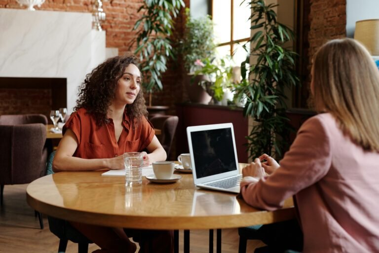 Two businesswomen engage in a meeting at a cozy coffee shop, discussing work on a laptop.