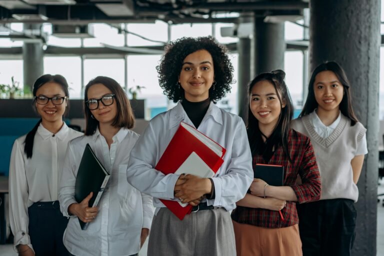 Group of diverse women colleagues smiling in a modern office setting, holding documents.
